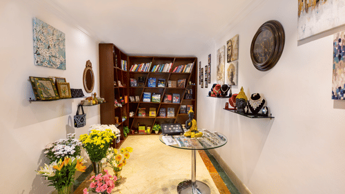 Souvenir shop at The Bristol Hotel, Gurgaon with a wooden bookshelf, framed artwork, a wall clock, jewellery displays, a Buddha statue, and flower bouquets.