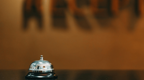 An image of a hand bell placed on a marble counter, with blurred, wall-mounted acrylic letters in the background.
