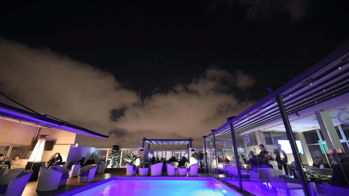 A luxurious, open-air lounge and swimming pool area at night, illuminated by numerous lights under a covered structure.