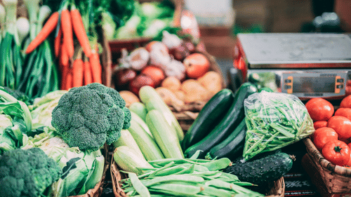 A display of fresh vegetables including broccoli, carrots, beans, tomatoes, and leafy greens arranged on a market stall.