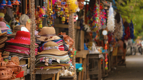A street market with stalls displaying hats, bags, and textiles with people walking past.