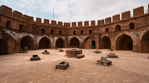 An image of a large fort courtyard with arched walls surrounding and a few stone structures inside.
