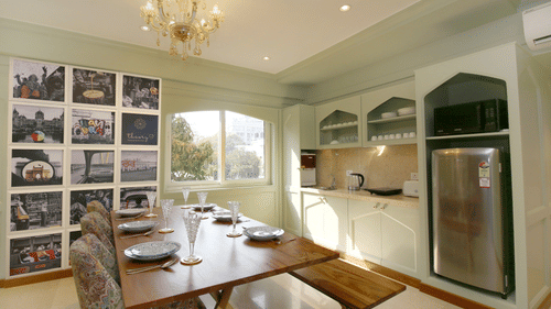 A kitchen with a table and chairs on one side, a wooden bench on the other, a collage of hotel images on the wall, and a window providing a view outside