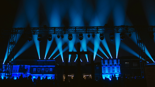 A stage with multiple beams of light in different direction under the night sky and an excited audience