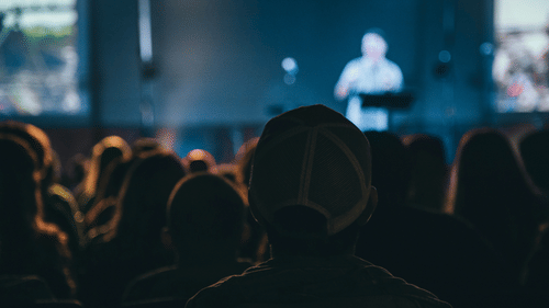A view from the back of a conference where many people are listening to the speaker on stage.