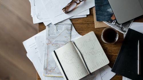 A cluttered wooden desk with papers, a notebook, glasses, a laptop, a drink, and other miscellaneous items