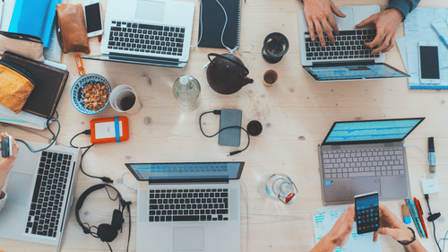A table with five laptops, phones, notebooks, coffee cups, and hands working on devices