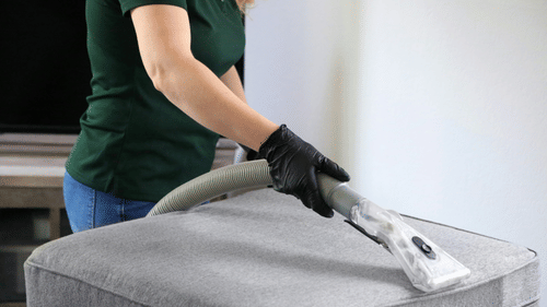 A person vacuuming a gray upholstered bench with a handheld attachment.