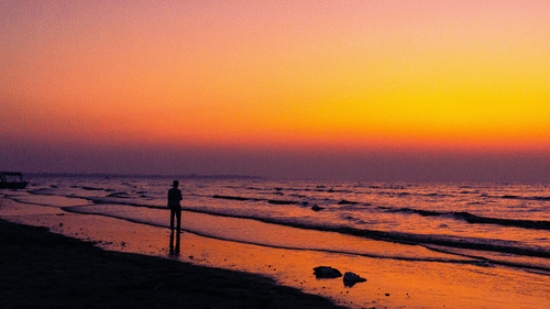 Vibrant sunset over Daman Beach with gentle waves, colourful skies, and a visitor standing along the water's edge