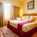 Hotel room at Featherlite Evoma Hotel with a bed dressed in white and maroon linen, wooden furnishings and long curtains by the window nearby.