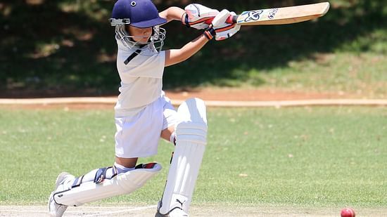A young cricketer wearing protective gear plays a shot on a green field, with the red ball bouncing on the pitch nearby.