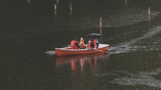 A small orange rowing boat carrying 3 people gliding across the calm surface of a dark lake.