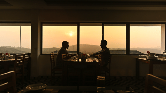Two people sit at a dining table silhouetted against a large window showing a sunset over hilly terrain | Abad Silvermist, Vagamon