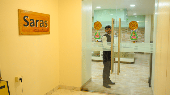 An entrance to Saras restaurant at Ambar Sarovar Portico, Gandhidham features a glass door, a staff member, and a wooden signboard on the wall.