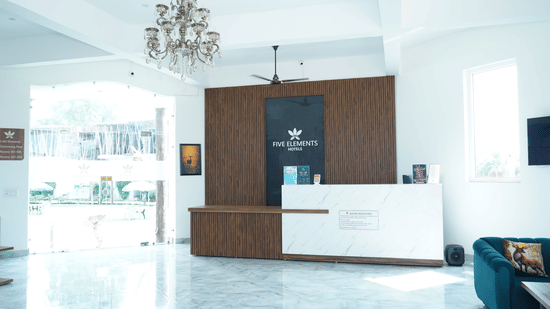 Reception area featuring a modern front desk counter with branding, wood paneling background, crystal chandelier, and bright tiled floors - Five Elements Hotels, Corbett Crown Resort, Jim Corbett
