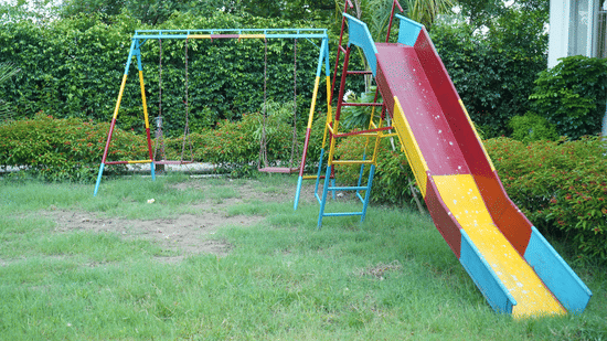 Children's outdoor playground area on a grass lawn featuring a green swing set and a red, yellow, and blue plastic slide - Five Elements Hotels, Corbett Crown Resort, Jim Corbett