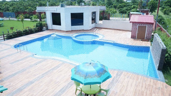 Elevated high-angle view of the outdoor swimming pool area showing the crystal blue water, paved patio area, and a blue patio table umbrella - Five Elements Hotels, Corbett Crown Resort, Jim Corbett