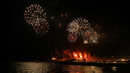 Fireworks explode in the night sky above a body of water reflecting the lights of a nearby dock or shore.