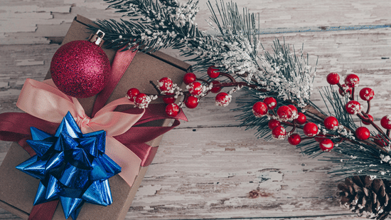 A rustic wooden surface displays festive Christmas decorations including a snowy branch, red baubles, and a wrapped gift with a blue bow.