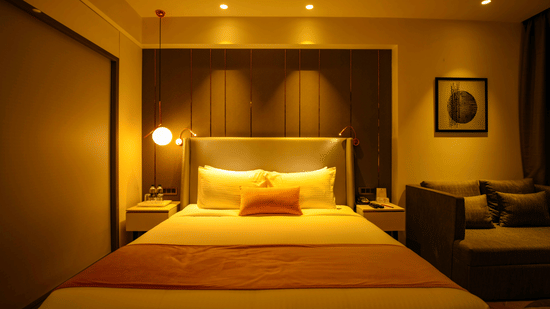 A luxurious hotel room with a plush king-sized bed, golden lighting, bedside lamps, and a cosy seating area with a sofa - Hotel Kanha Shyam, Prayagraj