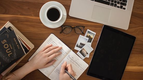 an image of a laptop, coffee cup, glasses and a woman writing something inside a notebook on a wooden table