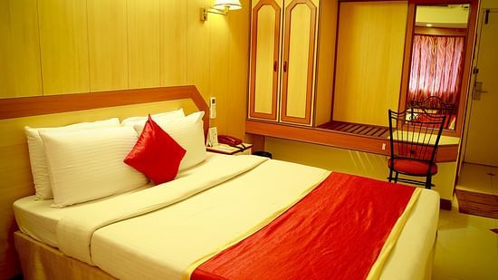 a room displaying a bed covered in white bed sheet and red pillow with a wardrobe and looking glass placed by its side - Hotel Swagath, Bangalore