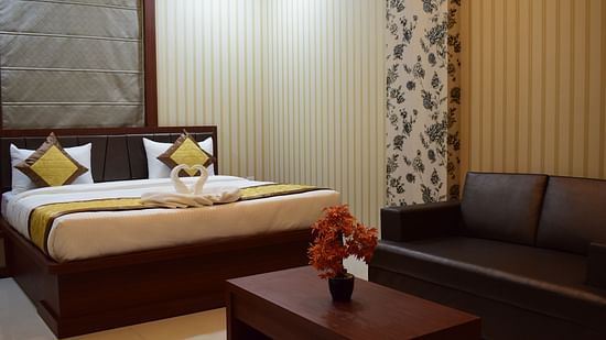 A spacious room at Hotel Uday Palace featuring a large bed, a cushioned sofa, and a wooden table with a flower vase.