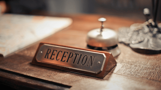 A close-up shot of a classic brass 'Reception' sign and a service bell on a wooden hotel front desk.