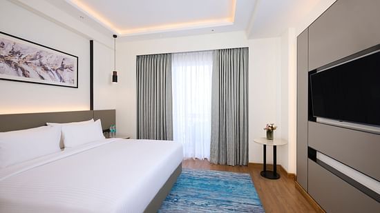 A comfortable hotel bedroom includes a large bed with white linens, a blue rug on the wooden floor, a wall-mounted television, and curtains partially drawn over the window. - Iris Sarovar Portico, Sector 85, Gurugram