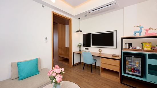 A welcoming hotel room is furnished with a sofa featuring a turquoise cushion, a desk with a blue chair, a wall-mounted television, a mini-fridge, and a small table with flowers. - Iris Sarovar Portico, Sector 85, Gurugram