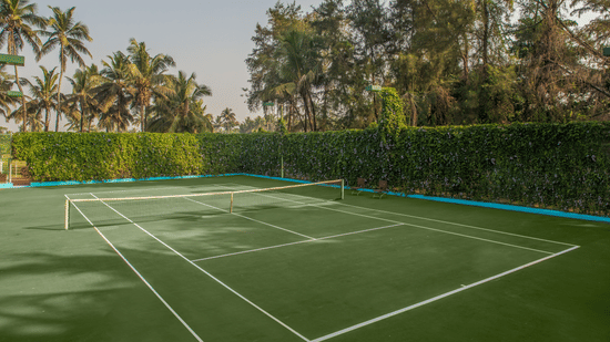 Green outdoor tennis court lined with hedge fencing and tall trees at Kenilworth Resort & Spa, Goa.