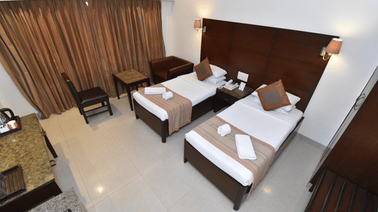 A Deluxe Room with 2 neatly-made beds, an A/C unit, a sitting area and full-length curtains | Kings International Juhu