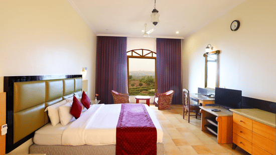 A bright hotel bedroom with a big bed, a desk and a TV, and 2 chairs by an arched window overlooking the greenery outside | Kohinoor Samudra Beach Resort, Ratnagiri