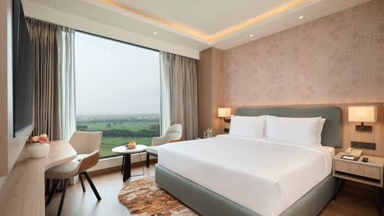 A bedroom at KPG Sarovar Portico, Raj Nagar Extension, Ghaziabad featuring a king size bed, a desk with chair, a window with outdoor views, and bedside tables with lamps.