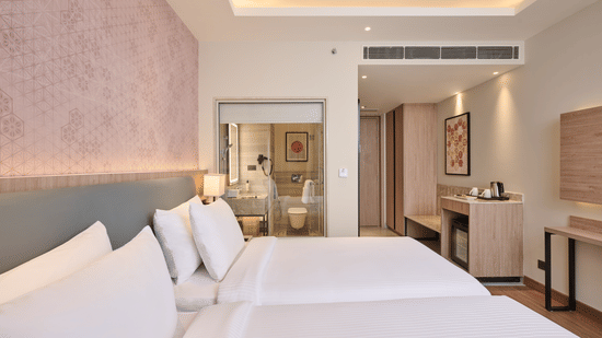 A bedroom at KPG Sarovar Portico, Raj Nagar Extension, Ghaziabad featuring twin beds, a tea station with a kettle, an entrance door, and a glass wall viewing the bathroom.