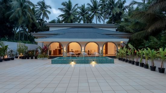 An exterior view of a white, low-rise villa with a swimming pool surrounded by a tiled patio and tropical greenery under a dusk sky - Lamrin Morjim Goa