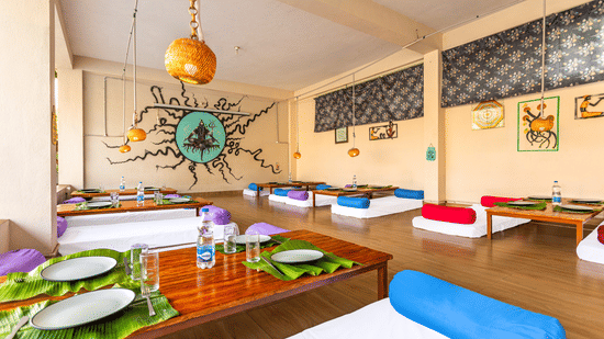 Low-angle view of a restaurant table set with banana leaves and steel plates, featuring colorful bolster cushions - Nature Trails Ashoka Resort Hampi