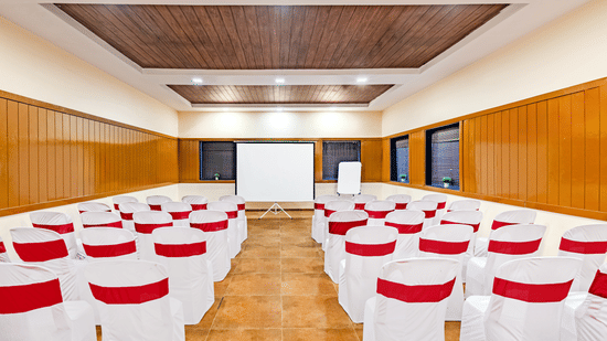 Nature Trails Kudalika conference hall with theatre seating, white and red chairs, projection screen, flipchart, wooden panelling and recessed ceiling lights.