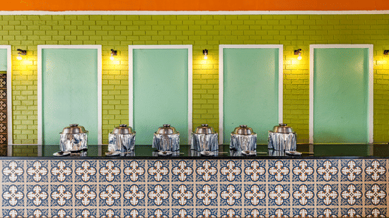 Nature Trails Kudalika dining buffet counter with steel chafing dishes on black granite top, blue patterned tile fascia, green brick wall and warm wall lights.