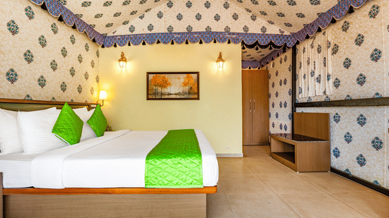 Nature Trails Kudalika premium AC tent room with double bed, green bedding, block-print fabric walls, brass wall lanterns, wooden wardrobe and framed wall painting.