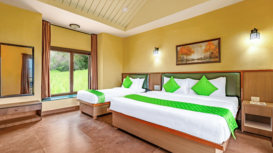 Nature Trails Kudalika river view room with two beds, green bedding, yellow walls, green headboards, wall lights, framed painting and large window with greenery view.