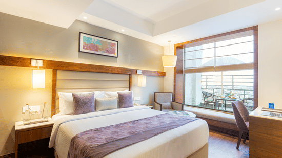 A bedroom at Nirvana Sarovar Portico, Gorakhpur featuring a king-size bed, bedside lamps, work station, and window overlooking a balcony.