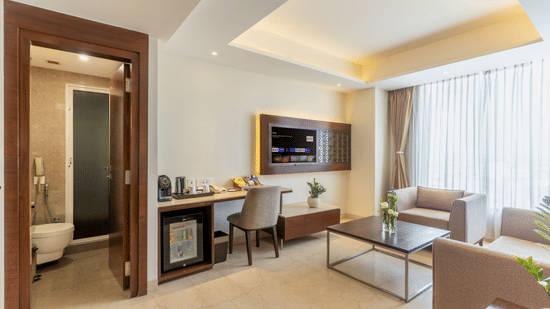 A hotel suite living area at Nirvana Sarovar Portico, Gorakhpur featuring a couch, work desk, wall-mounted television, and bathroom access.