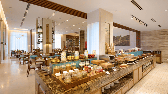Buffet setup at Prachi Sarovar Premiere, Bhubaneswar, showing a long stone counter with food items, dining tables, and overhead lighting fixtures.