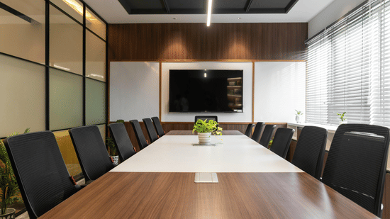 A professional conference room featuring a long, polished wood table with a center inlay. Dark ergonomic mesh chairs line both sides of the table. At the far end, a large flat-screen TV is mounted on a wood-paneled wall, flanked by acoustic panels. The roo