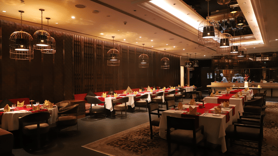 Restaurant at Pride Plaza Hotel, New Delhi featuring several sets of tables with chairs and lights hanging from the ceiling.