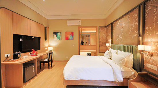 A premium guest room at Pride Premier Alwar featuring a large bed, a sleek entertainment unit with a mini-fridge, and colorful abstract wall art.