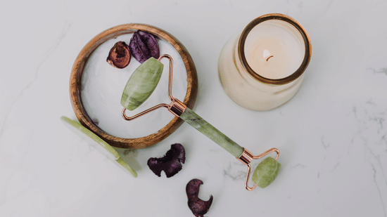 A jade roller lies on a white surface next to a lit candle and dried petals.