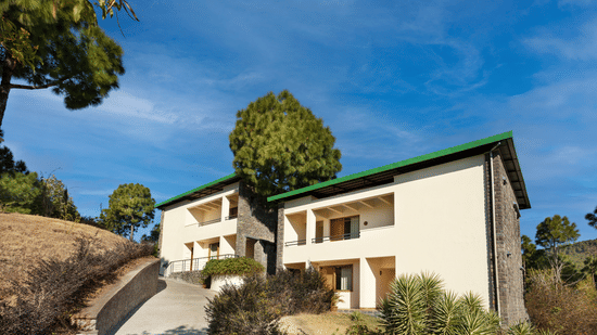 Exterior of premium rooms at Suryavilas Luxury Resort and Spa in Solan featuring the facade of the property on the mountain surrounded by trees and shrubs under a blue sky on a sunny day