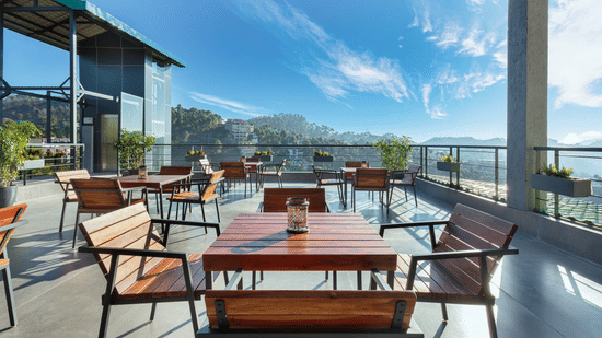 Outdoor dining area at Suryavilas Luxury Resort and Spa, featuring wooden tables and chairs on a spacious terrace with panoramic views of the Himalayan hills under a bright blue sky.